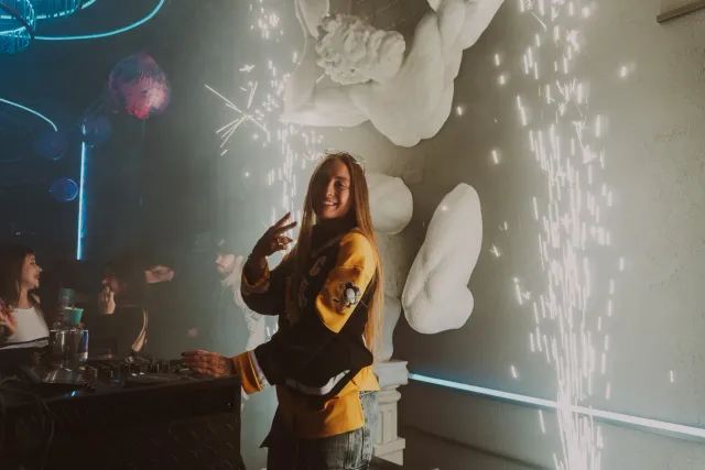 Chica posando junto a la cabina del DJ mientras una fuente de chispas ilumina la pared de esculturas blancas