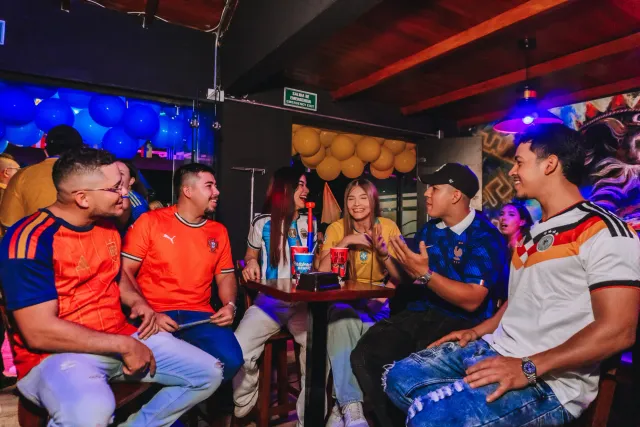 Grupo de amigos con camisetas de varias selecciones sentados alrededor de una mesa con vasos de Hermanos Drinks