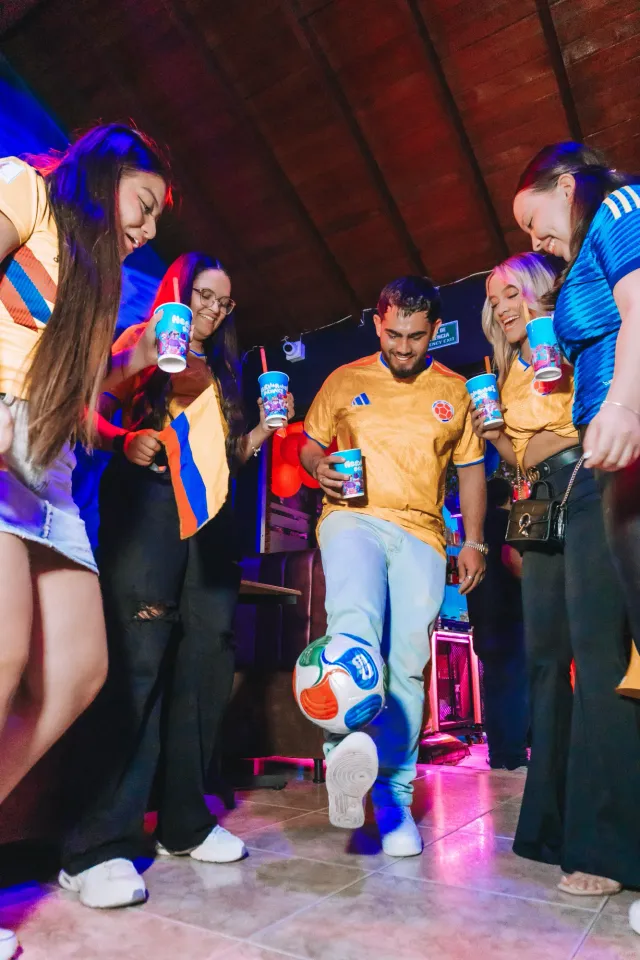 Amigos con camisetas de Colombia rodean a uno que domina un balón en el piso, todos con vasos en la mano